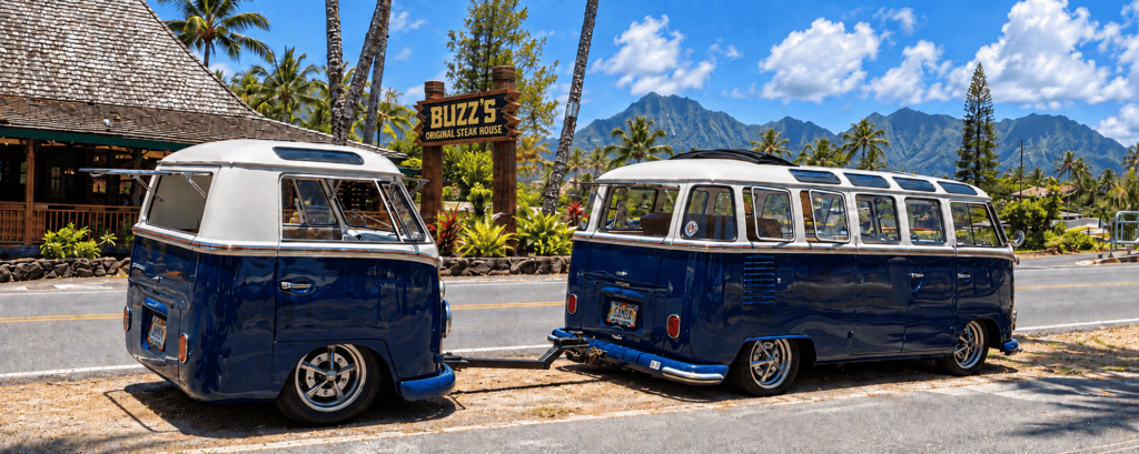 Two vintage blue-and-white VW buses beside Buzz’s Original Steak House in Kailua, with the restaurant sign and Koʻolau Mountains behind them